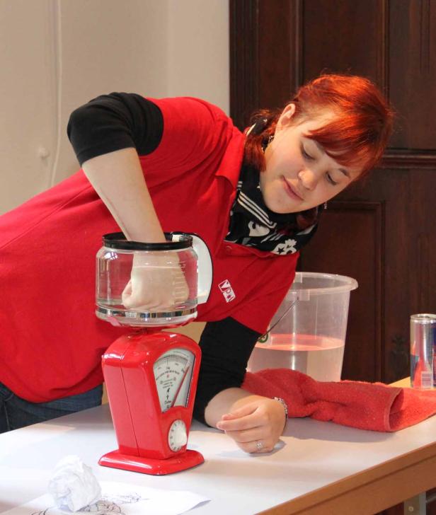 Eine Frau taucht ihre Hand in ein mit Wasser gefülltes Gefäß, das auf einer roten Waage steht.