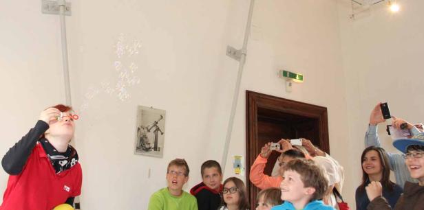 Eine Frau pustet Seifenblasen, während eine Gruppe Kinder und Erwachsene zuschaut und Fotos macht.