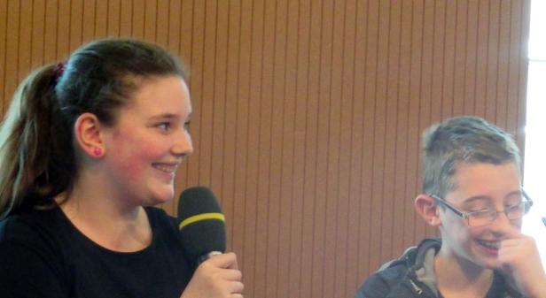 Ein Mädchen spricht in ein Mikrofon, während ein Junge daneben sitzt und lächelt.