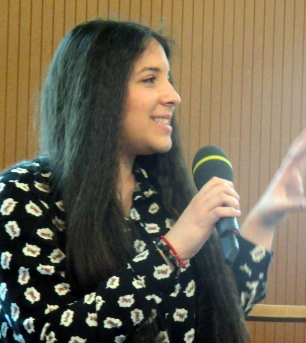 Eine Frau spricht in ein Mikrofon vor einem hölzernen Hintergrund.