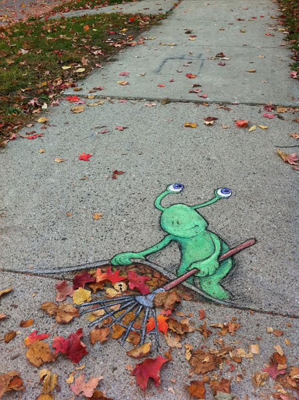 Ein grünes Alien mit Kulleraugen kehrt bunte Herbstblätter auf einem Gehweg.