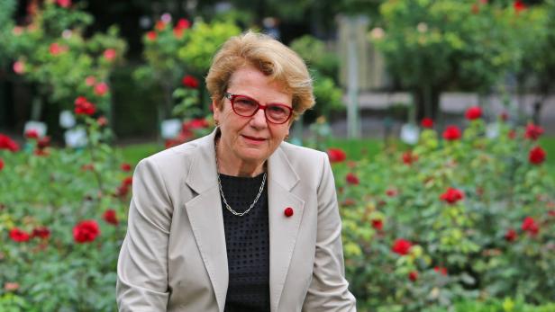 Eine ältere Frau mit Brille und Blazer posiert vor einem Rosenbeet.
