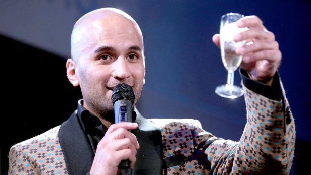Ein Mann mit Mikrofon in der Hand erhebt ein Glas Sekt.