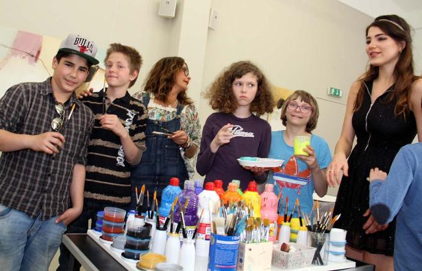 Eine Gruppe von Kindern und eine Frau stehen vor einem Tisch mit Farben und Pinseln.