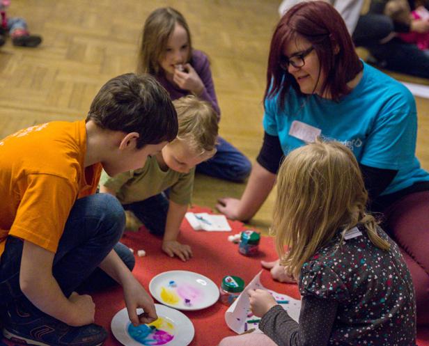 Eine Gruppe von Kindern sitzt mit einer Betreuerin auf dem Boden und bastelt mit Farben.
