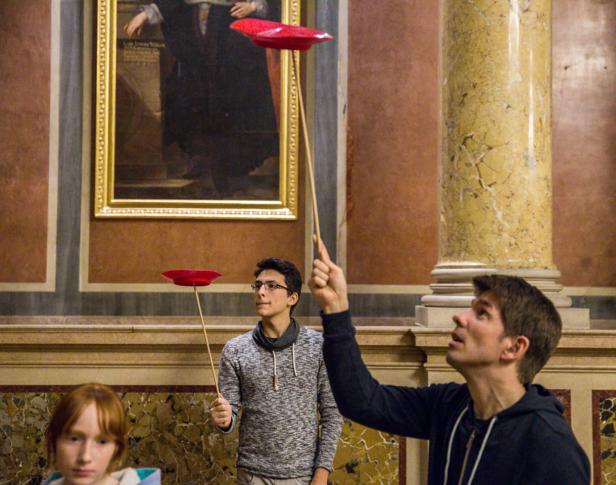 Mehrere Personen balancieren rote Teller auf Stäben in einem verzierten Raum.