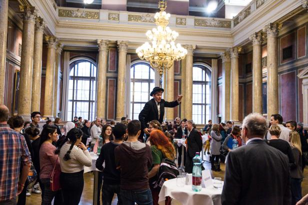 Ein Mann in einem Anzug steht auf Stelzen und spricht vor einer Menschenmenge in einem Ballsaal.