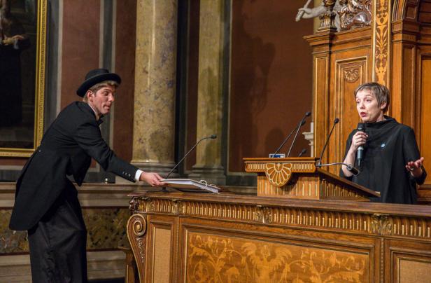 Eine Frau spricht an einem Rednerpult, während ein Mann ihr ein Dokument reicht.