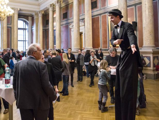 Ein Stelzenläufer im Anzug interagiert mit einer jungen Besucherin in einem großen Saal.