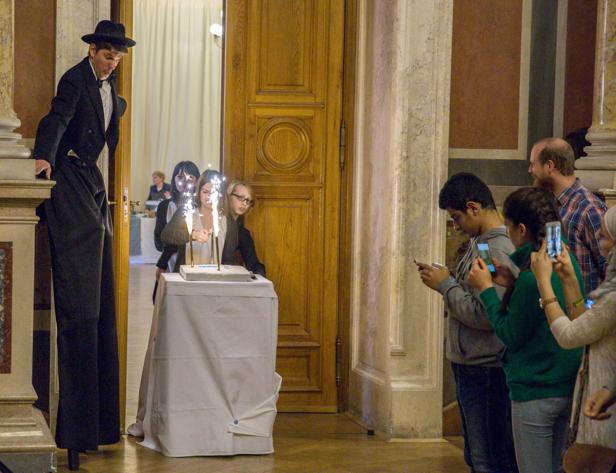 Ein Stelzenläufer steht neben einer Gruppe von Menschen, die ein Geburtstagstorte mit Wunderkerzen fotografieren.
