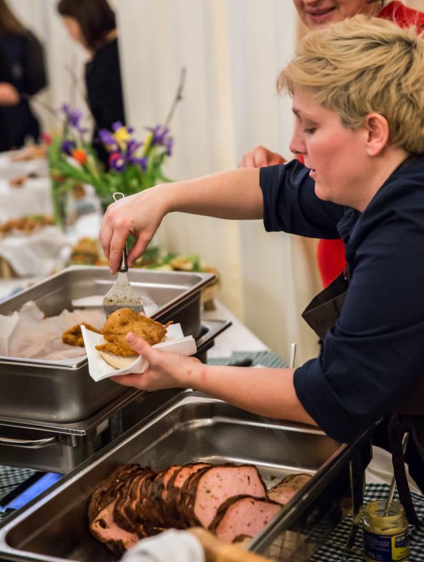 Eine Person serviert Essen von einem Buffet, darunter Fleischkäse und frittierte Speisen.