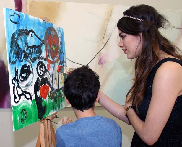 Eine Frau hilft einem Kind beim Malen eines bunten Bildes auf einer Staffelei.