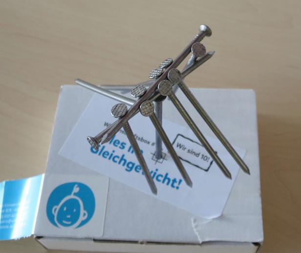 Mehrere Nägel balancieren auf einer weißen Schachtel mit blauem Logo.