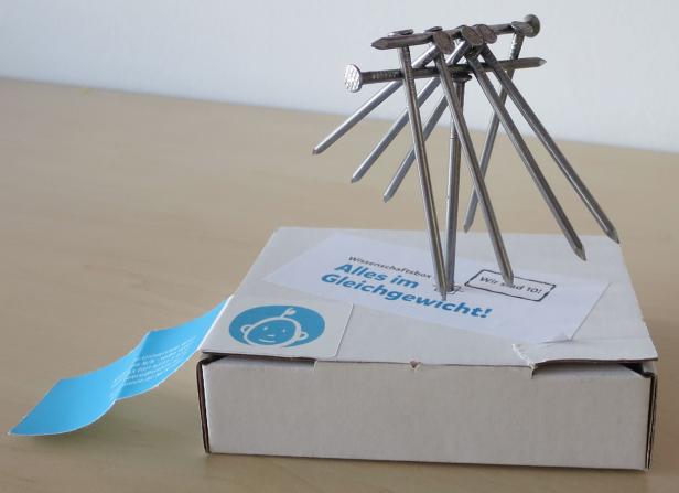 Mehrere Nägel balancieren auf einer Schachtel mit der Aufschrift „Alles im Gleichgewicht!“.