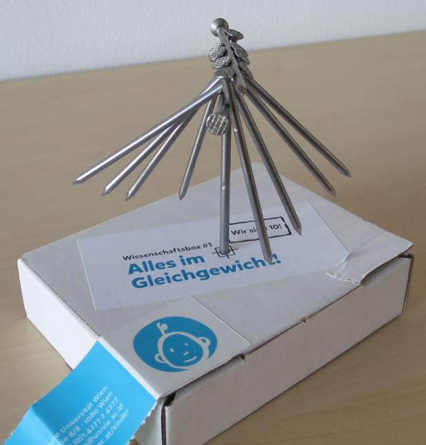 Ein Nagel-Mobile steht auf einer Schachtel der „Wissenschaftsbox #1: Alles im Gleichgewicht!“.