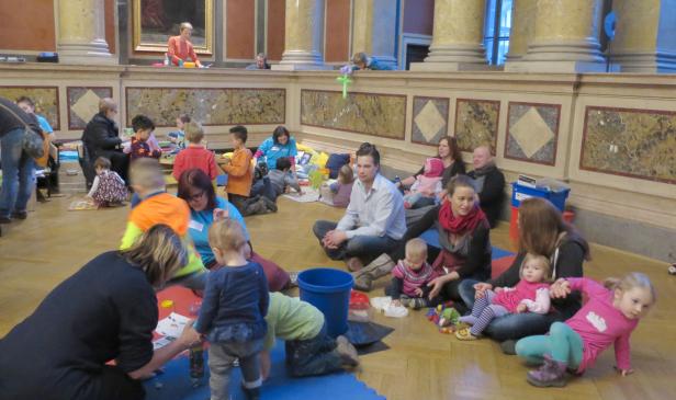 In einem Raum mit Säulen spielen Kinder mit ihren Eltern auf dem Boden.