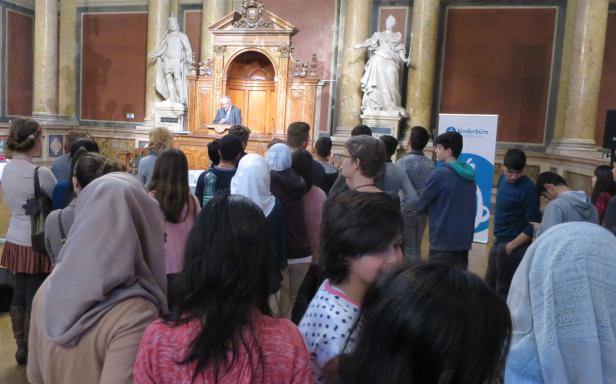 Eine Gruppe von Menschen hört einem Redner in einem verzierten Saal zu.