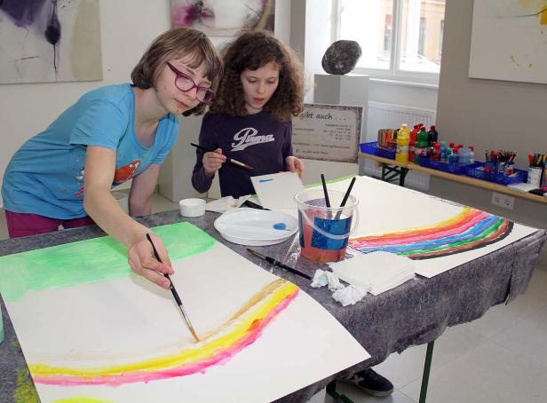 Zwei Mädchen malen einen Regenbogen auf Papier in einem hellen Raum.