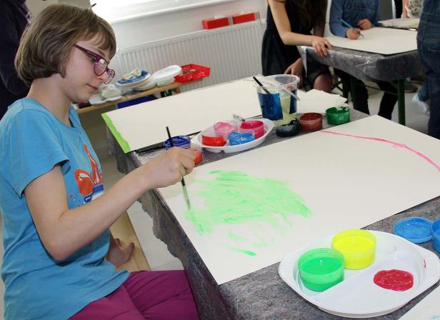 Ein Mädchen mit Brille malt mit grüner Farbe auf ein großes Blatt Papier.