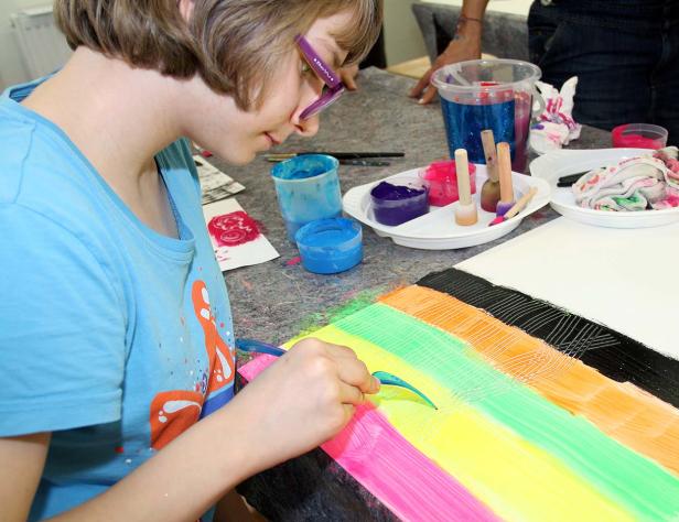 Ein Mädchen kratzt mit einem Werkzeug Farbe von einem regenbogenfarbigen Bild.