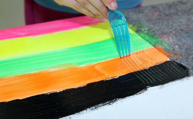 Eine Person zieht mit einer Gabel Linien durch nasse, bunte Farbe auf einer Leinwand.