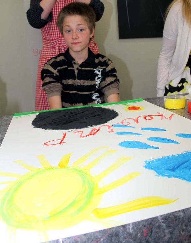 Ein Junge steht vor einem bemalten Plakat mit Sonne, Wolken und einem schwarzen Loch.