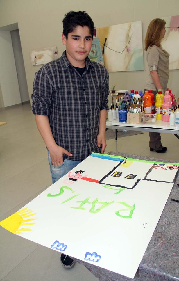 Ein Junge steht vor einem Tisch mit einem bunten Kinderbild von Häusern und der Aufschrift „RATIS“.