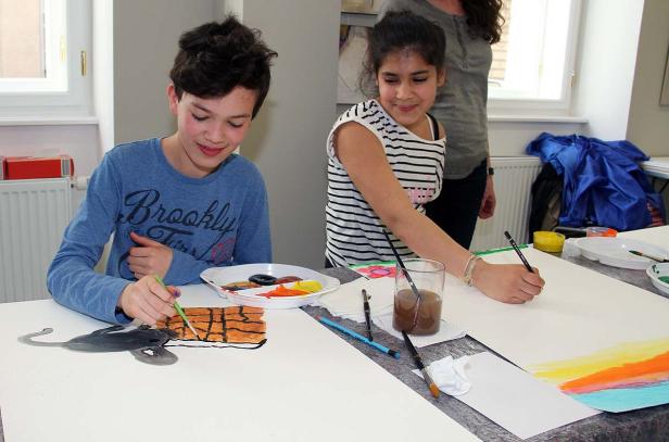 Zwei Kinder malen an einem Tisch mit Pinseln und Farben.