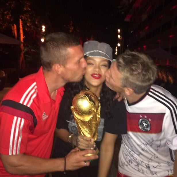 Rihanna hält die FIFA-Weltmeisterschaft und wird von zwei deutschen Fußballspielern auf die Wange geküsst.