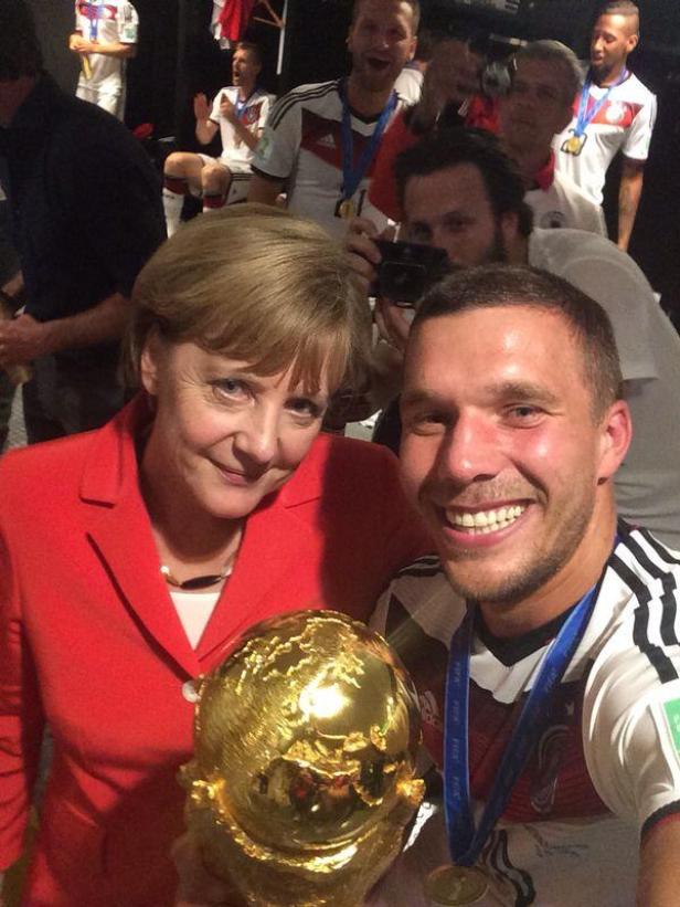 Angela Merkel posiert mit Lukas Podolski und dem WM-Pokal, umringt von weiteren deutschen Nationalspielern.