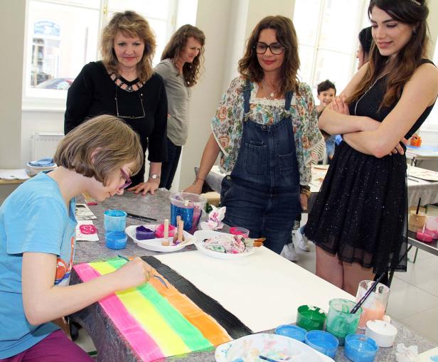 Ein junges Mädchen malt einen Regenbogen auf ein Blatt Papier, umgeben von anderen Personen.