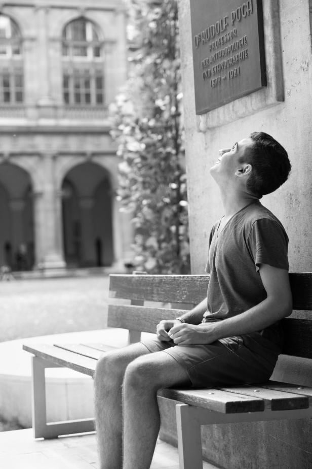Ein junger Mann sitzt auf einer Bank und schaut zu einem Schild von D. Rudolf Poch hinauf.