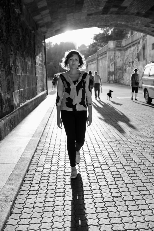 Eine Frau geht auf einem gepflasterten Weg entlang, mit anderen Personen und einem Hund im Hintergrund.