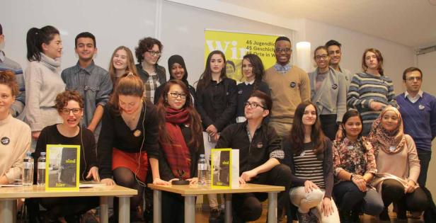 Eine Gruppe junger Menschen posiert vor einem Banner mit der Aufschrift „Wir sind hier“.