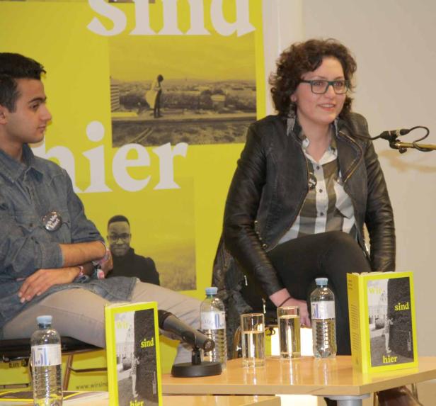 Eine Frau mit Brille und ein Mann sitzen auf einer Bühne vor einem gelben Banner.
