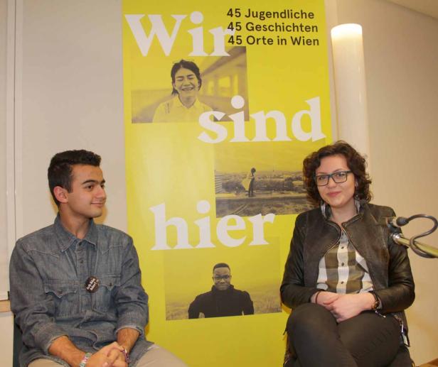 Ein junger Mann und eine junge Frau sitzen vor einem Plakat mit dem Titel „Wir sind hier“.
