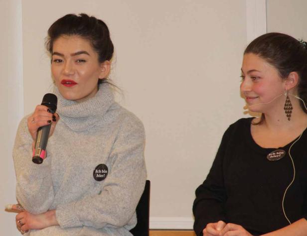 Zwei Frauen, beide mit Buttons „Ich bin hier!“, sprechen vor Publikum.