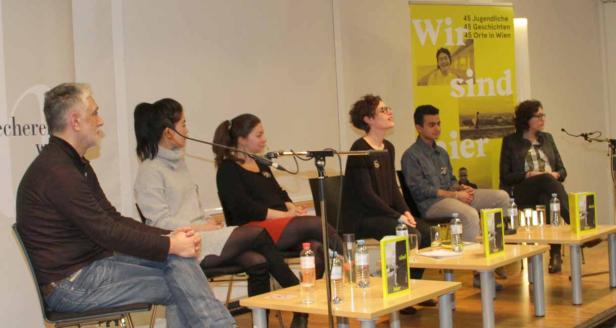 Sechs Personen sitzen an Tischen auf einer Bühne während einer Podiumsdiskussion.