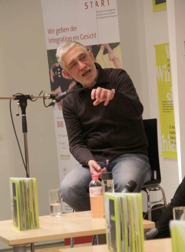 Ein Mann mit grauem Haar sitzt auf einem Stuhl und gestikuliert vor einem Mikrofon.