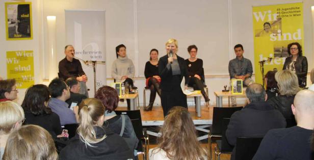 Eine Frau spricht bei einer Veranstaltung vor Publikum, im Hintergrund sitzen weitere Personen auf einer Bühne.