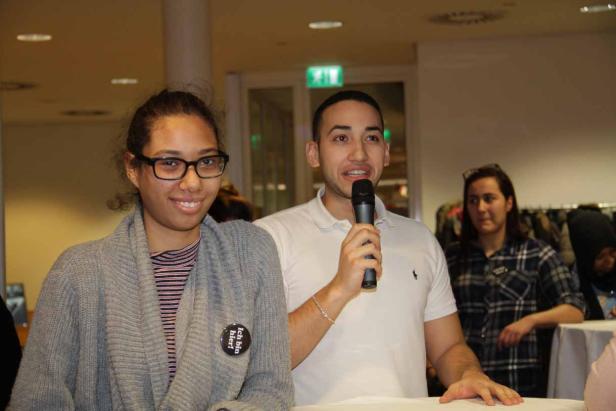 Ein junger Mann spricht bei einer Veranstaltung ins Mikrofon, neben ihm eine junge Frau mit einem Button „Ich bin hier!“.
