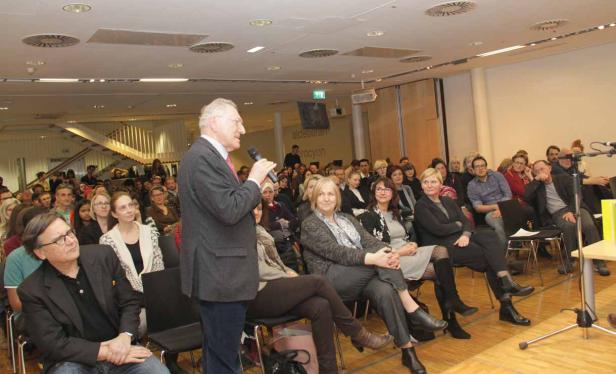 Ein Mann spricht mit Mikrofon vor einem Publikum in einem hellen Raum.