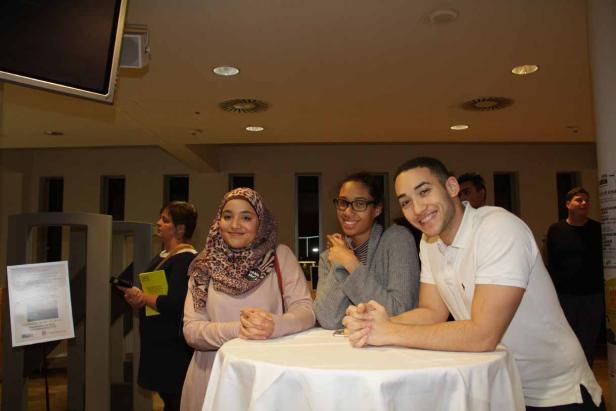 Drei junge Leute stehen lächelnd an einem Tisch bei einer Veranstaltung.