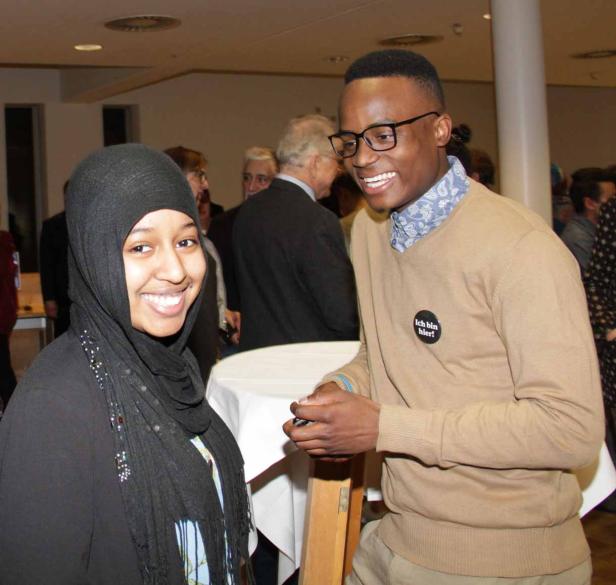 Eine junge Frau mit Hijab und ein junger Mann mit einem „Ich bin hier!“-Button lächeln.