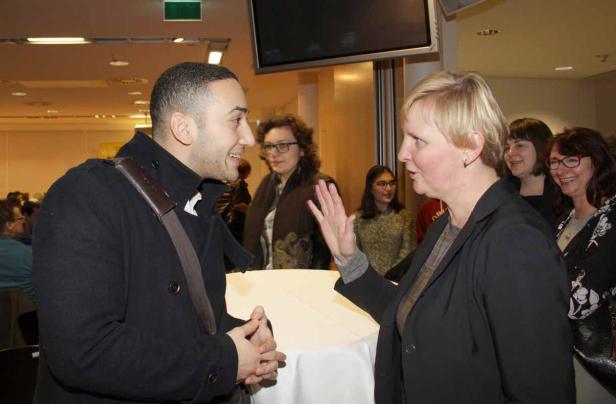 Eine Frau gestikuliert, während sie sich mit einem Mann unterhält; weitere Personen im Hintergrund.