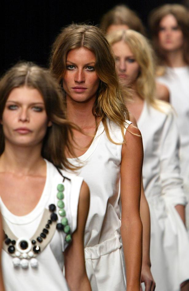Gisele Bündchen und andere Models in weißen Kleidern auf einem Laufsteg.