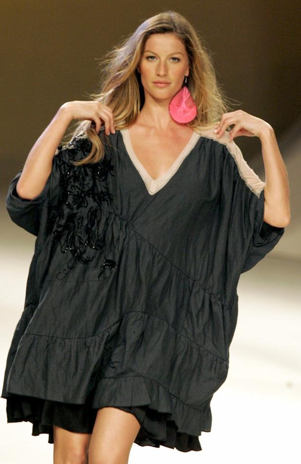 Gisele Bündchen präsentiert ein weites, dunkles Kleid mit Rüschen und einem pinkfarbenen Ohrring.