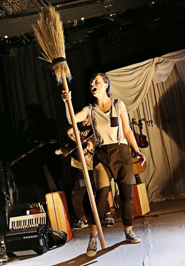 Eine Frau mit Hosenträgern hält einen Besen auf einer Bühne.