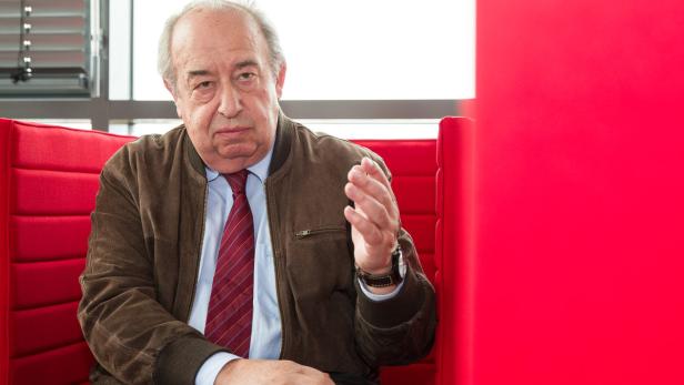 Ein Mann mit brauner Jacke sitzt in einem roten Sessel und gestikuliert.