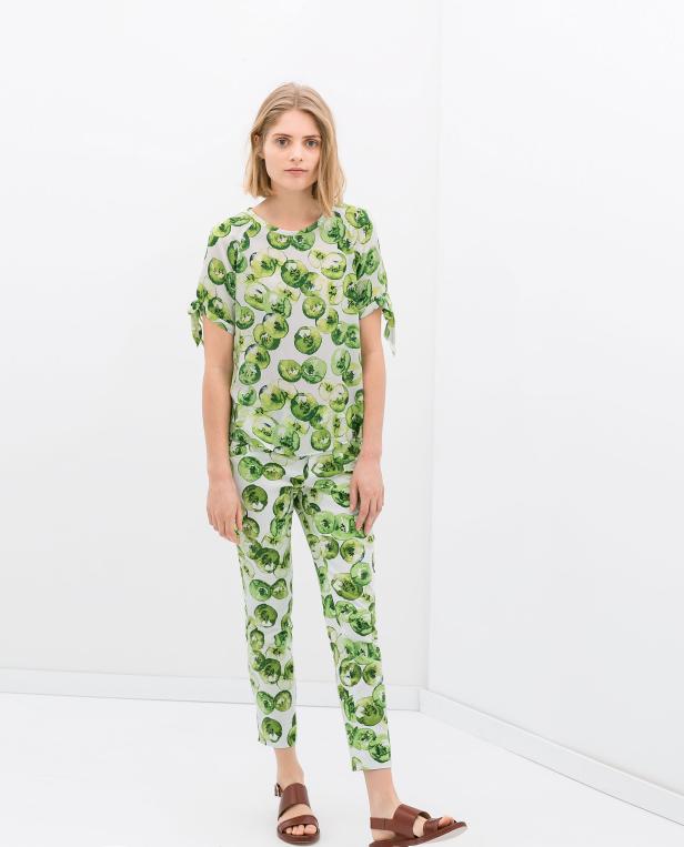 Eine Frau in einem passenden Outfit mit grünem Kiwi-Muster und braunen Sandalen steht in einem weißen Raum.